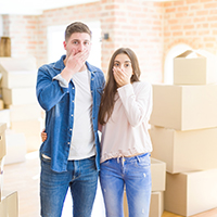 newly moved couple with hand covering their mouth with boxes in the background