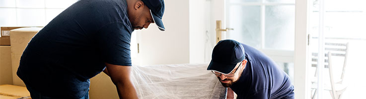 two men wrapping a piece of furniture in a room full of moving boxes