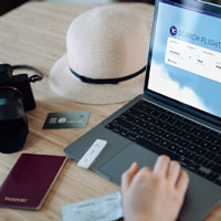 camera and hat beside a laptop with a cropped hand of someone shopping online for flight tickets on an airline website