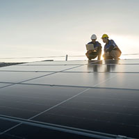 two engineers wearing hard hats looking at a laptop on a rooftop with solar panels