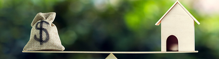 us dollar in sack bag and wooden house model on opposite ends of a scale with green tree bokeh as background
