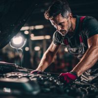 A car mechanic in overalls leaning over the engine compartment with work light positioned behind him