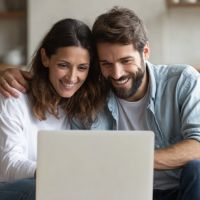a smiling man and a woman who seems to be a couple are sitting together in front of a laptop