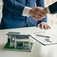 two men shaking hands with a model house and paperwork on a desk